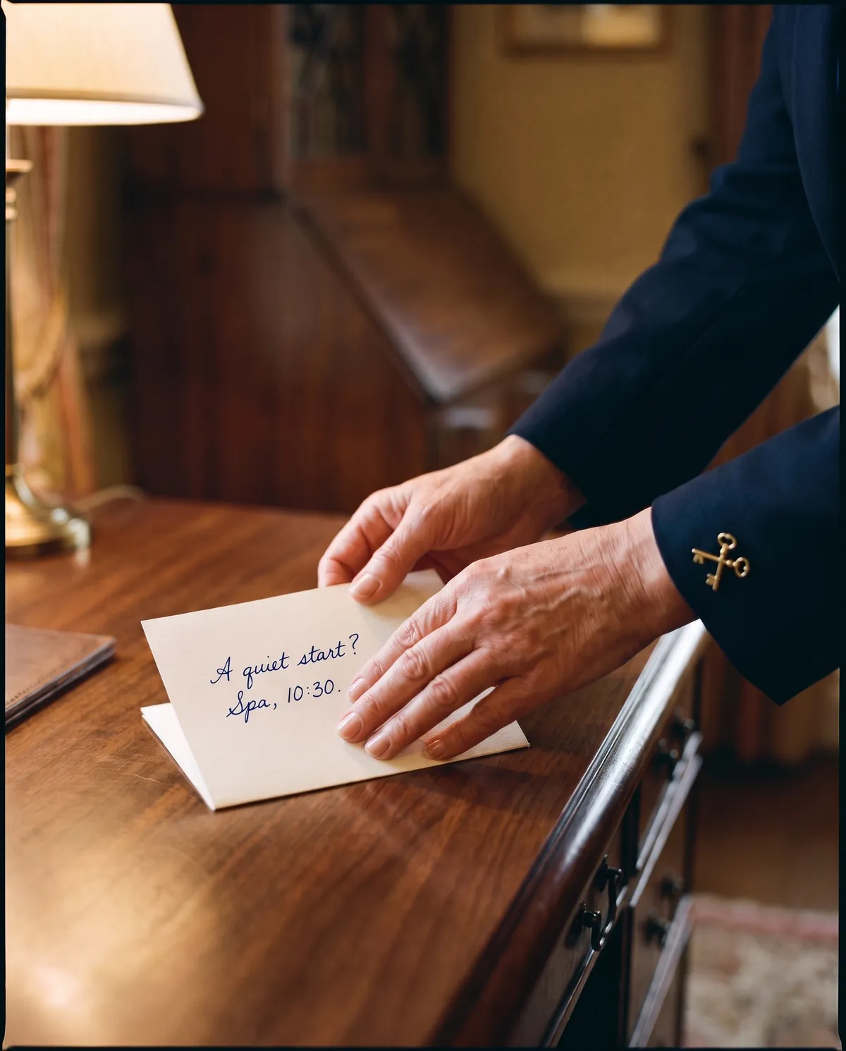 A concierge in a navy uniform with a golden crossed-keys pin places a folded card on a polished desk; handwritten on it: A quiet start? Spa, 10:30.