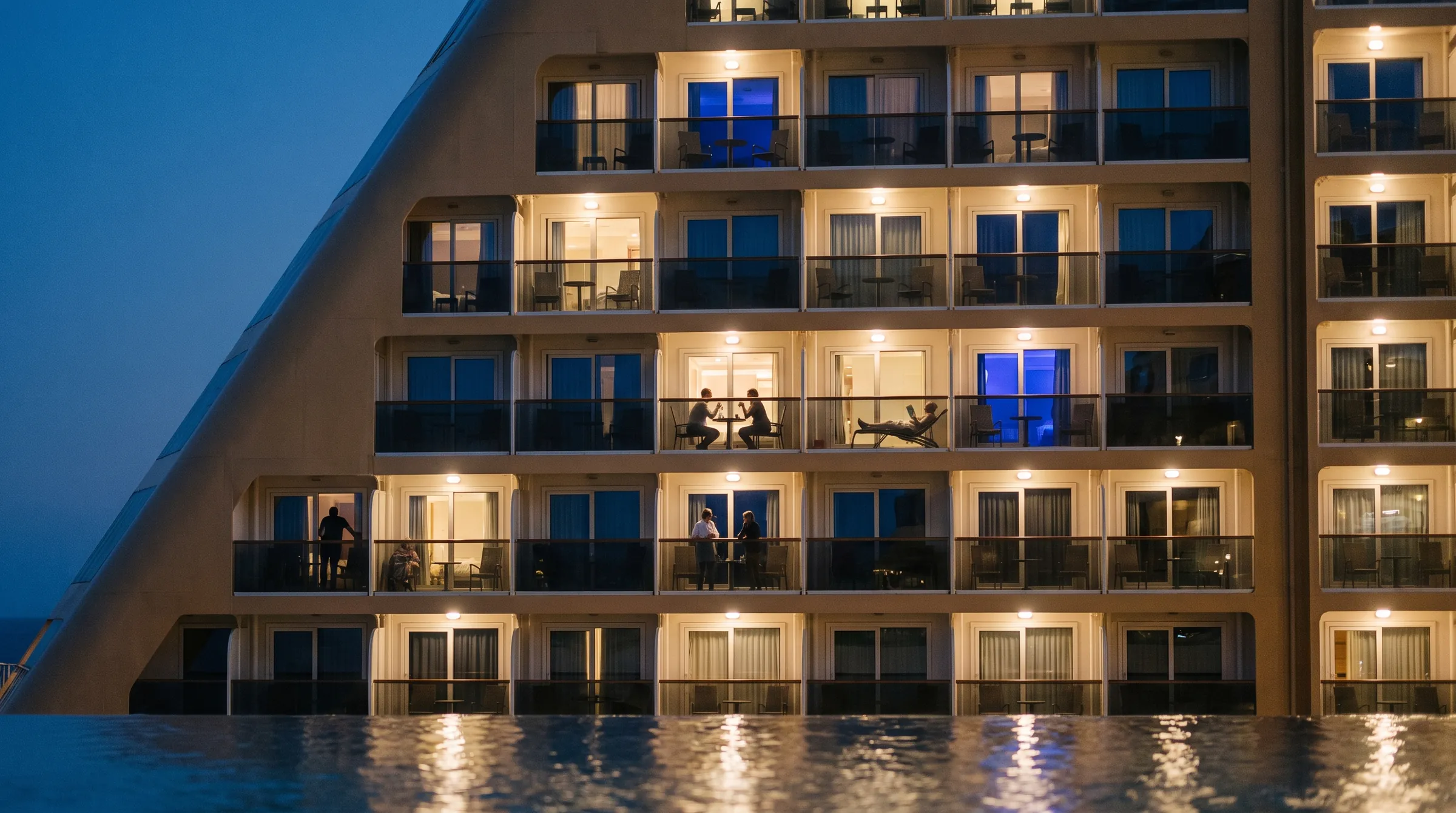 The balconies of a cruise ship at dusk, every cabin lit differently, a few guests out on their balconies